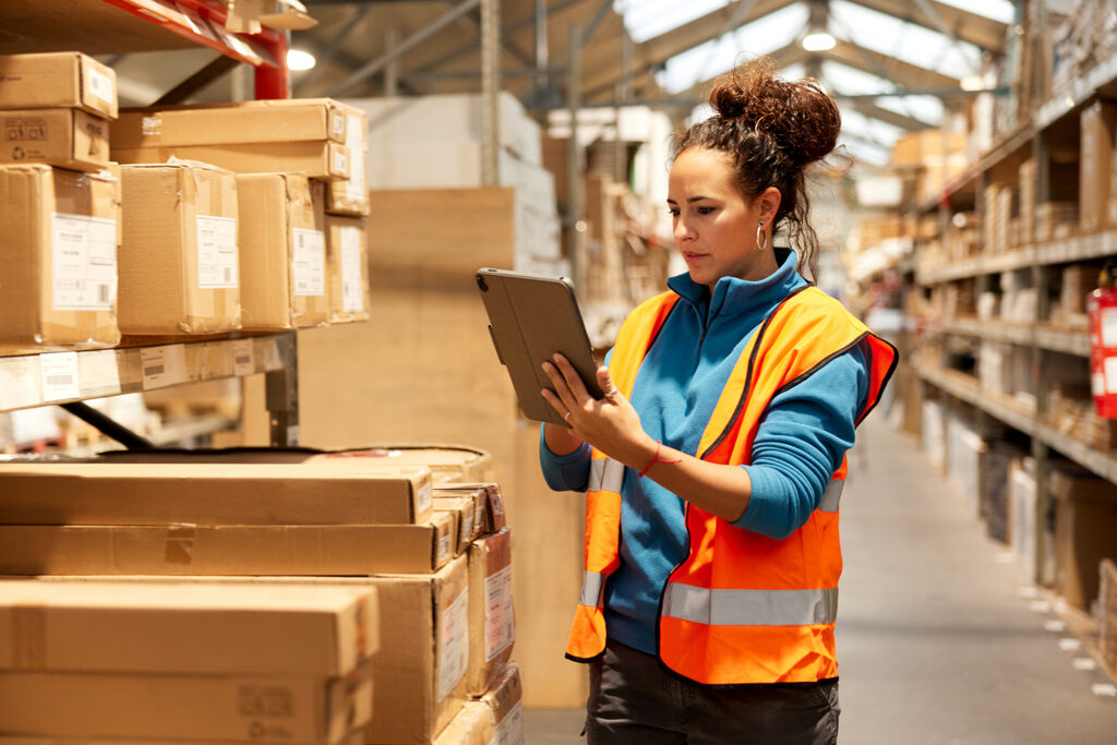 A warehouse worker is standing next to a shelf and using a digital tablet.